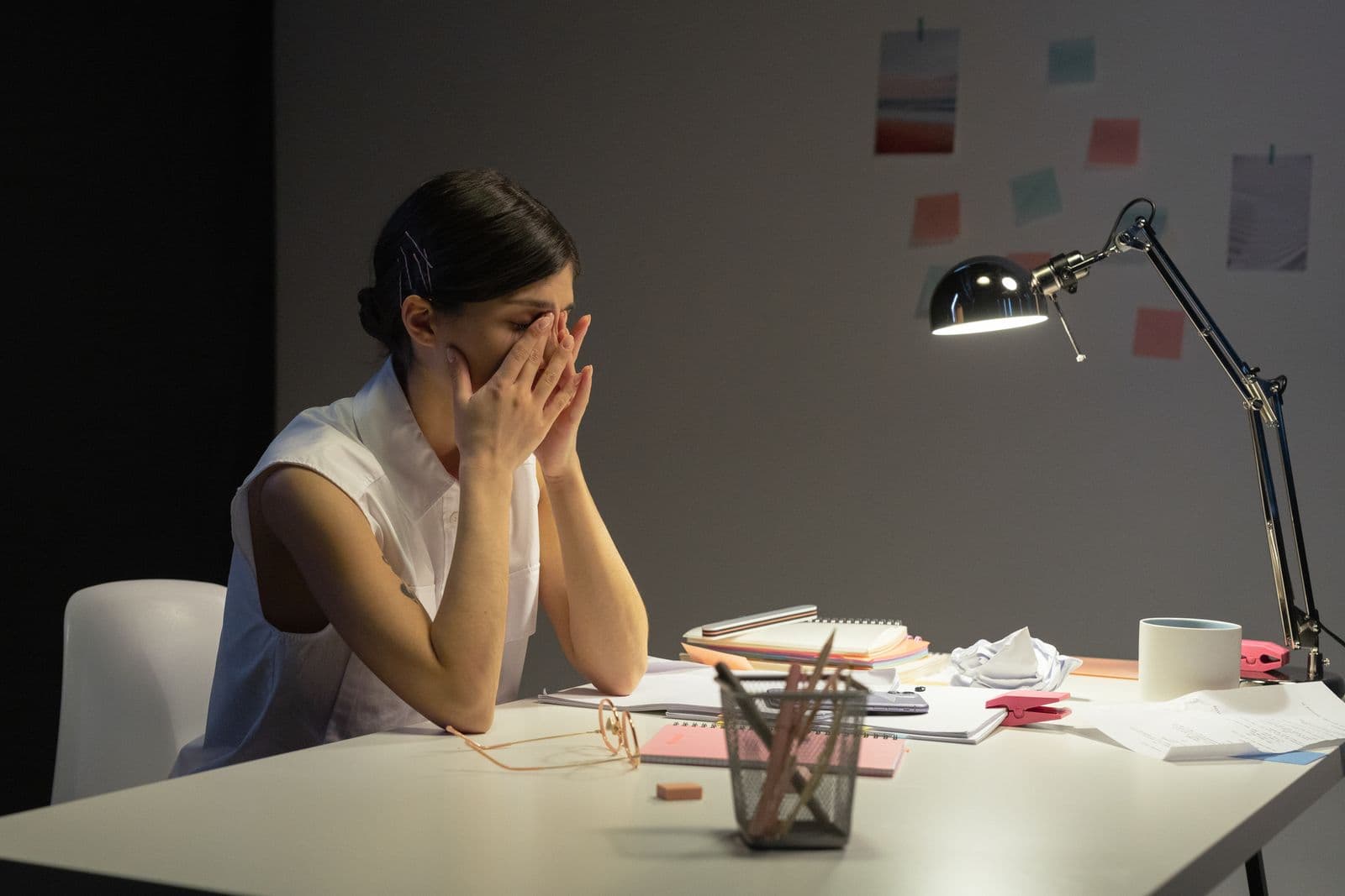 A tired woman rubbing her eyes at a cluttered desk late in the day